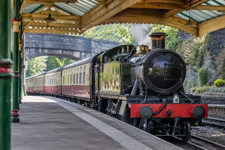 A preserved railway carriage passing beneath a station canopy with visible clearances viewed from a safe platform.