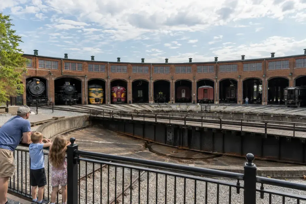 An illustrative brick railway roundhouse and turntable viewed from behind a railing.